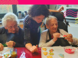 une animatrice se penche vers deux dames âgées qui font un atelier pâtisserie ; elles décorent des biscuits