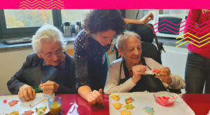 une animatrice se penche vers deux dames âgées qui font un atelier pâtisserie ; elles décorent des biscuits