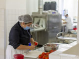 une dame, avec masque chirurgical et charlotte, prépare des tomates dans une cuisine professionnelle.