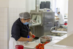 une dame, avec masque chirurgical et charlotte, prépare des tomates dans une cuisine professionnelle.