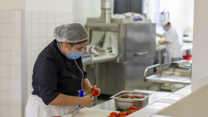 une dame, avec masque chirurgical et charlotte, prépare des tomates dans une cuisine professionnelle.