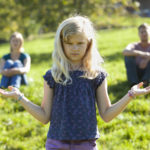 une jeune fille à l'air sérieux ouvre les paumes de ses mains comme dans un geste de choix difficile entre deux possibilités, au-dessus de ses mains on voit au loin, flous, un homme et une femme qui semblent être ses parents.