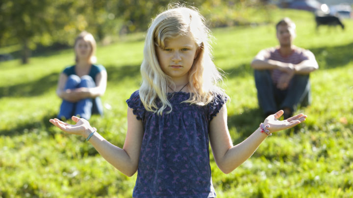 une jeune fille à l'air sérieux ouvre les paumes de ses mains comme dans un geste de choix difficile entre deux possibilités, au-dessus de ses mains on voit au loin, flous, un homme et une femme qui semblent être ses parents.