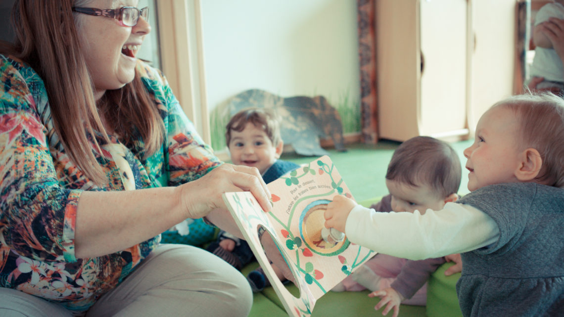 une dame montre un livre à un groupe de touts petits, l'un d'eux interagit avec elle et ils se sourient.