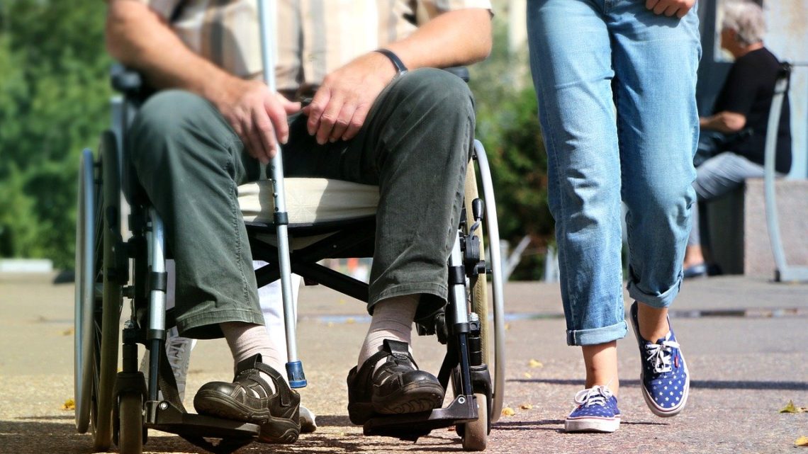 un homme en fauteuil roulant et une jeune personne marchant à côté (le cadrage ne montre que leurs jambes)