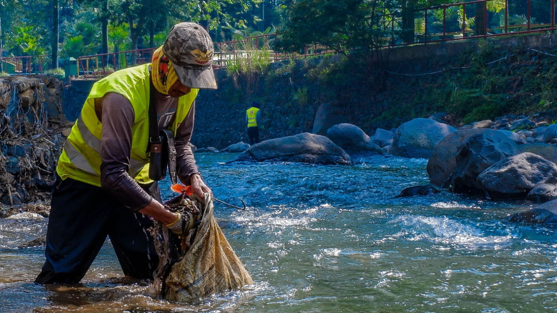 un homme avec gilet jaune, marche dans un fleuve/rivière avec un sac en tissu et semble mettre quelque chose dedans