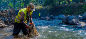 un homme avec gilet jaune, marche dans un fleuve/rivière avec un sac en tissu et semble mettre quelque chose dedans