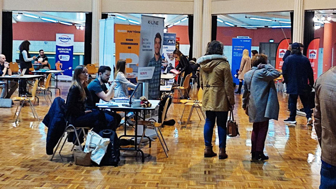 dans une grande salle, des stands d'entreprise et kakémonos sont installés, les candidats naviguent entre les tables pour les rencontrer et déposer leur cv.