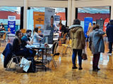 dans une grande salle, des stands d'entreprise et kakémonos sont installés, les candidats naviguent entre les tables pour les rencontrer et déposer leur cv.