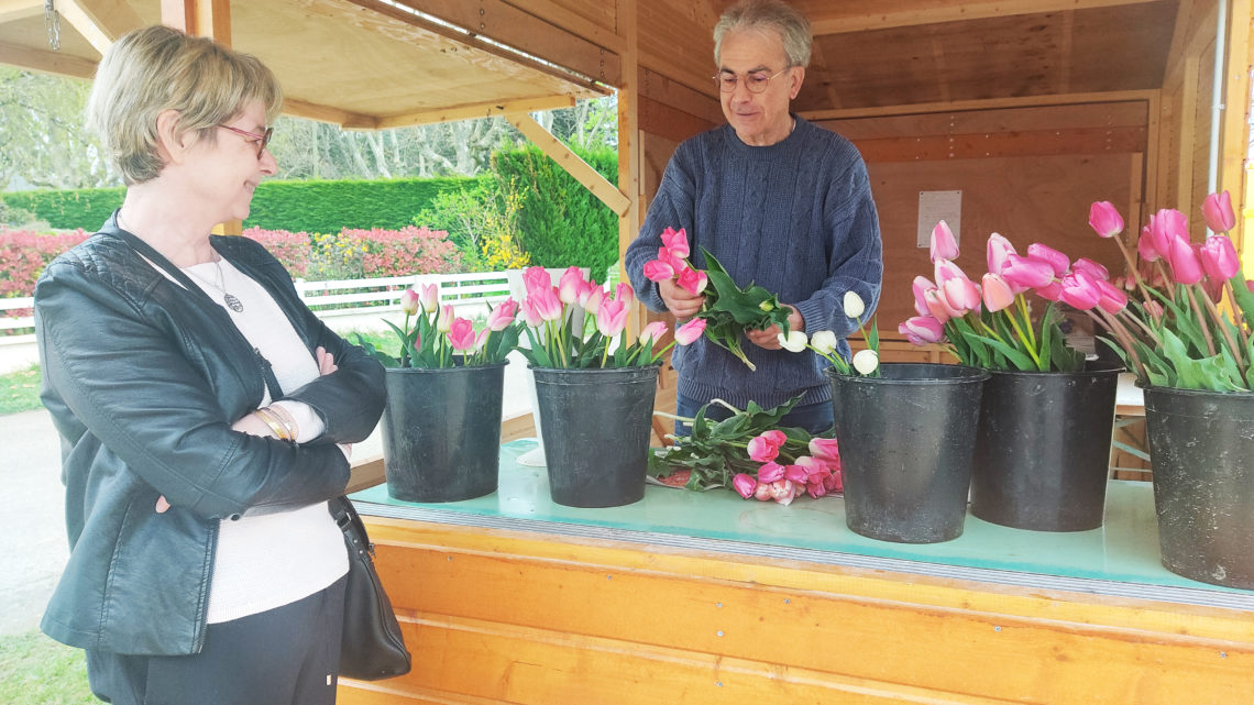 Une donatrice est en train de choisir son bouquet de tulipes sur un stand extérieur en bois, un homme s'occupe de les trier et d'encaisser les dons.