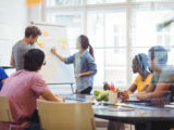 Des personnes en réunion travaillent et l'une d'elles écrit sur un paperboard.