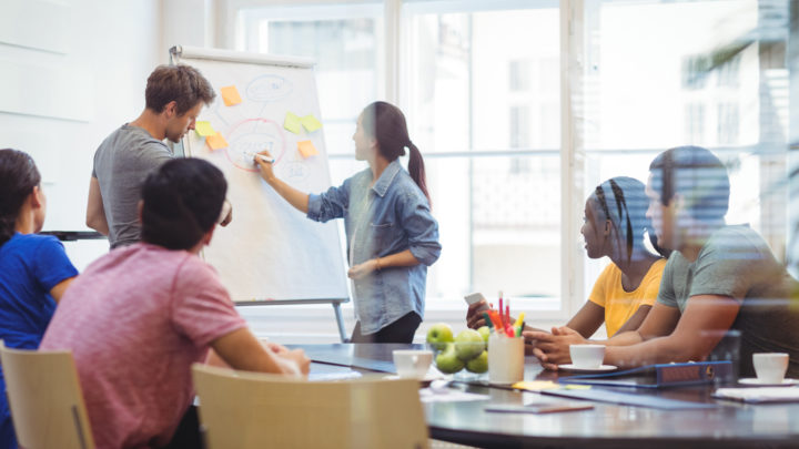 Des personnes en réunion travaillent et l'une d'elles écrit sur un paperboard.