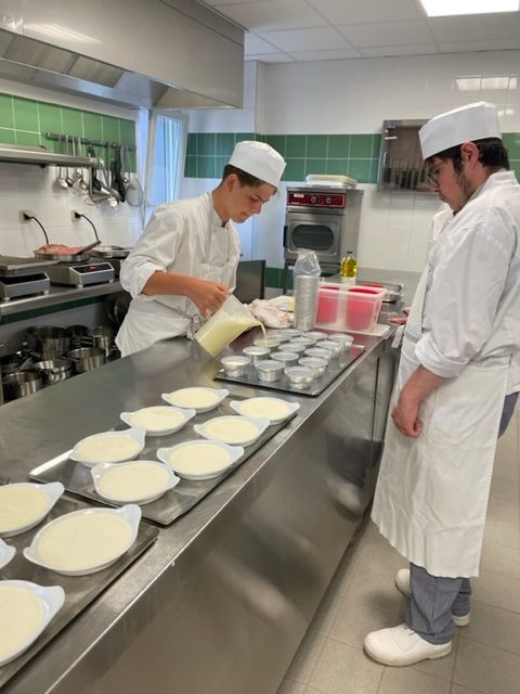 Devant un long plan de travail, deux jeunes préparent une tablée de verrines et moules, l'un d'eux verse une préparation ressemblant à une pâte à gâteau dans ces moules.