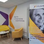 Locaux d'Xcelium : à l'intérieur, un bâtiment entièrement rénové et moderne avec un coin convivial (fauteuil, canapé,) et des éléments de décoration murale / roll-up aux couleurs de l'agence : violet et jaune.