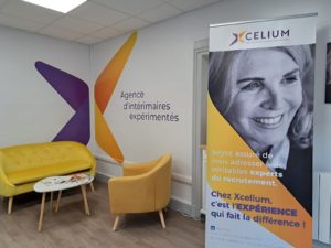 Locaux d'Xcelium : à l'intérieur, un bâtiment entièrement rénové et moderne avec un coin convivial (fauteuil, canapé,) et des éléments de décoration murale / roll-up aux couleurs de l'agence : violet et jaune.