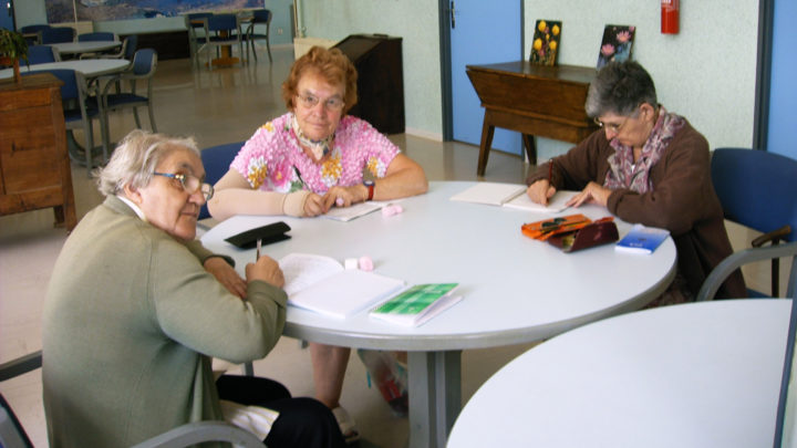 Trois dames sont autour d'une table, en train d'écrire sur leur carnet respectif.