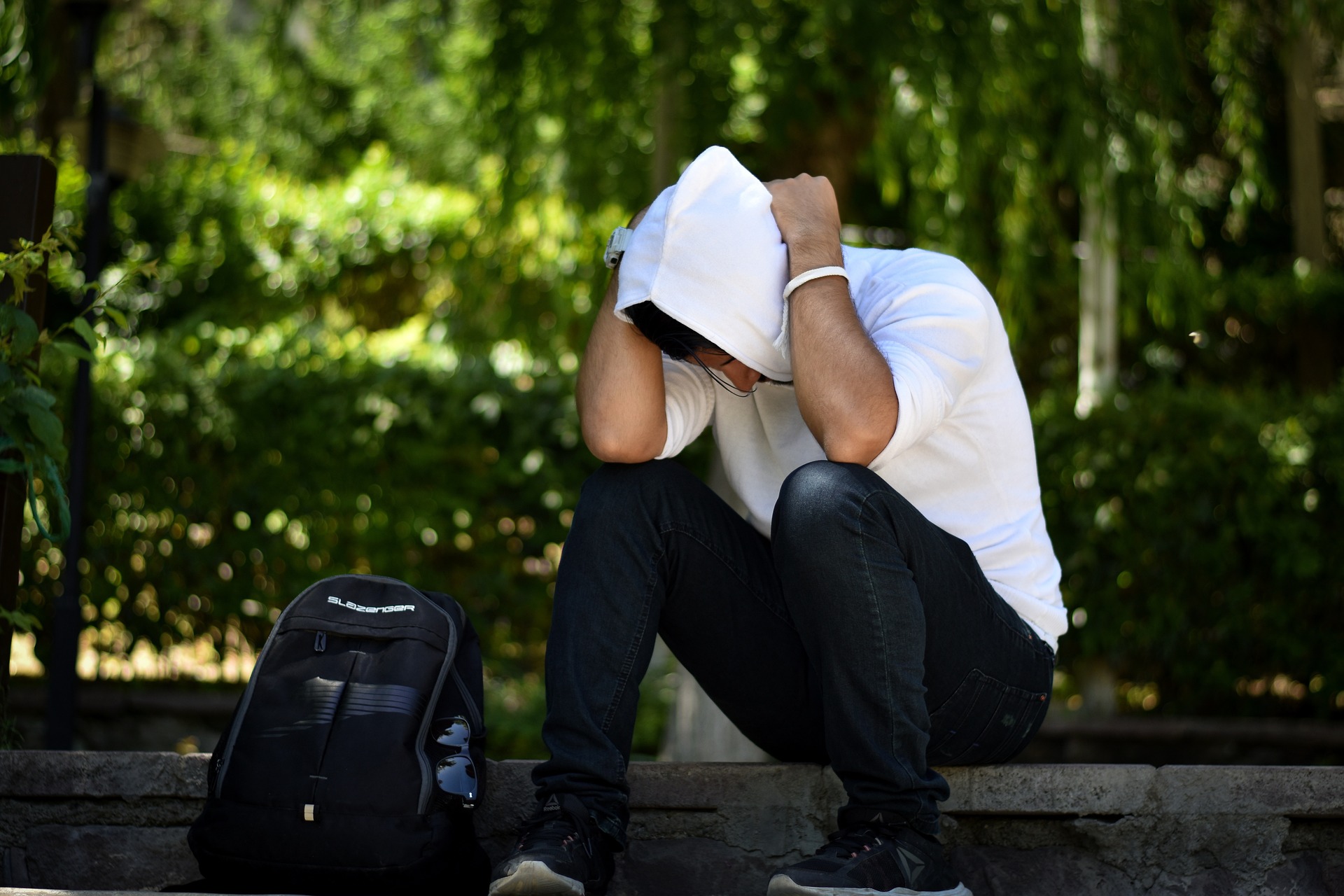 Sur un banc dans un parc, son sac à dos posé à côté de lui, un jeune homme en sweatshirt à capuche, se prend la tête entre les mains de façon préoccupée - photo de ©Hamed Mehrnik (Pixabay)