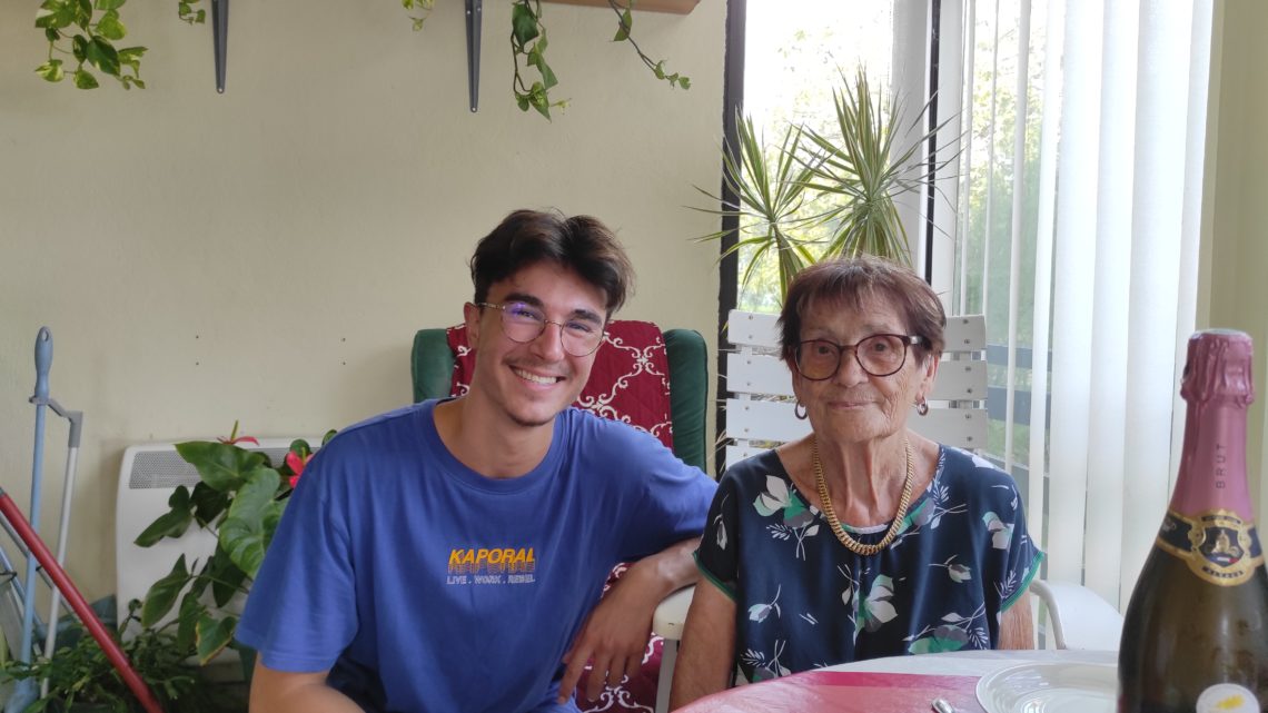 Résidence intergénérationnelle VEEC : un jeune homme et une dame âgée posent en souriant pour la photo, sur la table de leur salon, avec une bouteille de pétillant et de nombreuses plantes vertes d'intérieur.