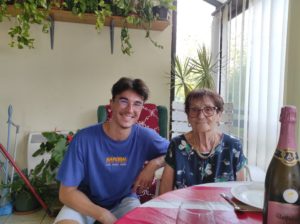 Résidence intergénérationnelle VEEC : un jeune homme et une dame âgée posent en souriant pour la photo, sur la table de leur salon, avec une bouteille de pétillant et de nombreuses plantes vertes d'intérieur.