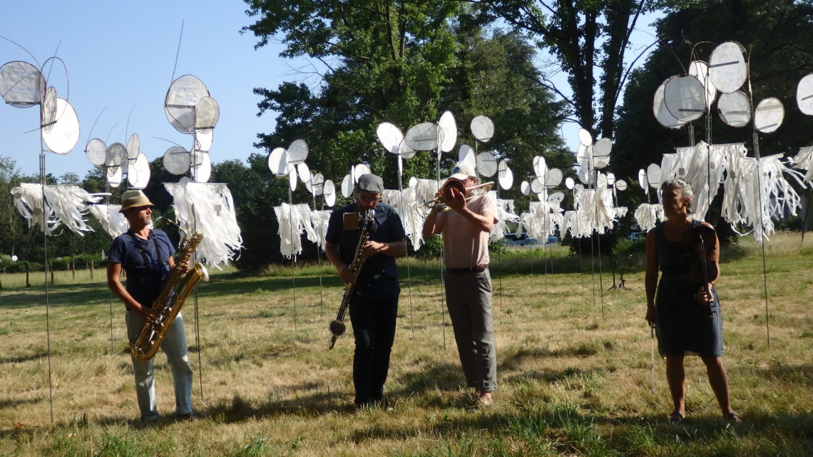 4 musiciens jouent de la trompette dans un champ devant des installations de l'expo "« D’où vient le vent… ». Derrière eux, les installations sont des girouettes, tissus attachés sur des piquets pour réagir au vent.