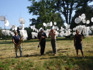 4 musiciens jouent de la trompette dans un champ devant des installations de l'expo "« D’où vient le vent… ». Derrière eux, les installations sont des girouettes, tissus attachés sur des piquets pour réagir au vent.