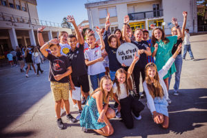 dans une cour de collège, un groupe d'enfants joyeux pose pour la photo avec un signe "Ici, c'est l'Ain"