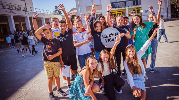 dans une cour de collège, un groupe d'enfants joyeux pose pour la photo avec un signe "Ici, c'est l'Ain"