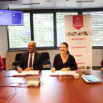 23 octobre 2023, sur une table devant une affiche de Grand Bourg Habitat, Isabelle Maistre (présidente – Grand Bourg Habitat) et Andy Nkundikije, (président – Mission Locale Bresse-Dombes-Côtière) signent la convention de partenariat pour répondre aux besoins de logement des jeunes et de colocation des seniors. Ils sourient au photographe. deux autres personnes sont assises à leurs côtés.