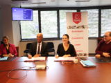 23 octobre 2023, sur une table devant une affiche de Grand Bourg Habitat, Isabelle Maistre (présidente – Grand Bourg Habitat) et Andy Nkundikije, (président – Mission Locale Bresse-Dombes-Côtière) signent la convention de partenariat pour répondre aux besoins de logement des jeunes et de colocation des seniors. Ils sourient au photographe. deux autres personnes sont assises à leurs côtés.