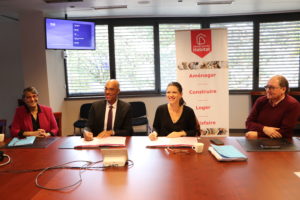 23 octobre 2023, sur une table devant une affiche de Grand Bourg Habitat, Isabelle Maistre (présidente – Grand Bourg Habitat) et Andy Nkundikije, (président – Mission Locale Bresse-Dombes-Côtière) signent la convention de partenariat pour répondre aux besoins de logement des jeunes et de colocation des seniors. Ils sourient au photographe. deux autres personnes sont assises à leurs côtés.