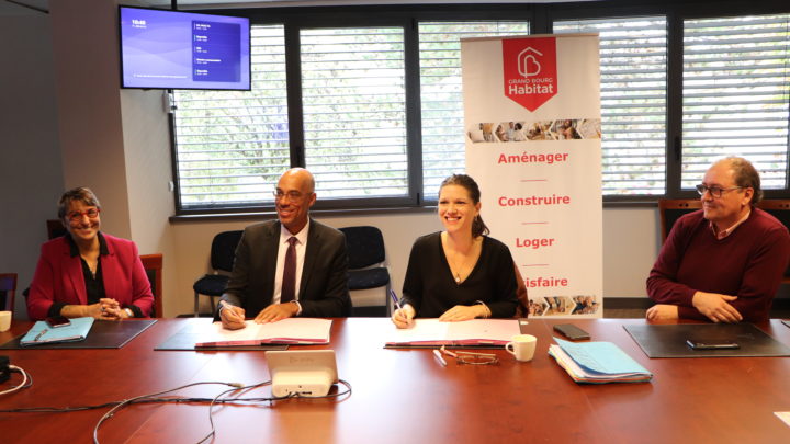 23 octobre 2023, sur une table devant une affiche de Grand Bourg Habitat, Isabelle Maistre (présidente – Grand Bourg Habitat) et Andy Nkundikije, (président – Mission Locale Bresse-Dombes-Côtière) signent la convention de partenariat pour répondre aux besoins de logement des jeunes et de colocation des seniors. Ils sourient au photographe. deux autres personnes sont assises à leurs côtés.