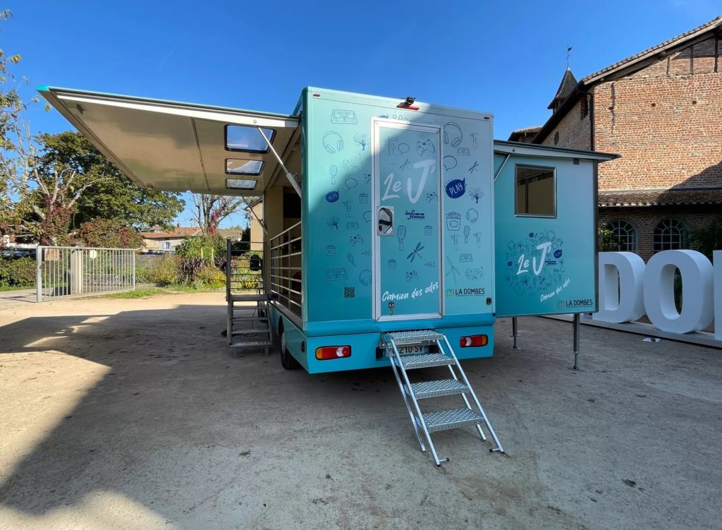 Initiative du réseau des animateurs jeunesse de la Communauté de communes de la Dombes, le camion Le « J » est un espace itinérant destiné aux 11-25 ans, Ici on voit le dos du camion, peint en bleu ciel, avec plein d'illustrations "jeunes" autour de la lettre "J" peinte en grand à l'arrière.