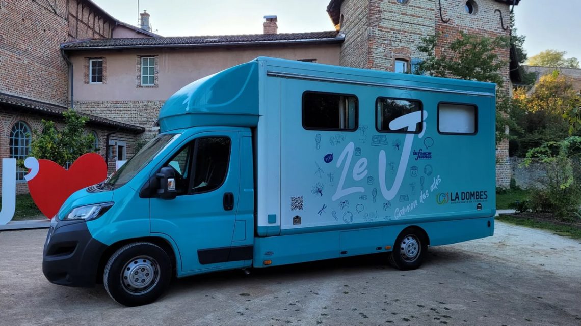 Initiative du réseau des animateurs jeunesse de la Communauté de communes de la Dombes, le camion Le « J » est un espace itinérant destiné aux 11-25 ans, Ici on voit le dos du camion, peint en bleu ciel, avec plein d'illustrations "jeunes" autour de la lettre "J" peinte en grand sur le côté du camion avec l'inscription "camion des ados" et le logo de la CC de la Dombes.