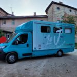Initiative du réseau des animateurs jeunesse de la Communauté de communes de la Dombes, le camion Le « J » est un espace itinérant destiné aux 11-25 ans, Ici on voit le dos du camion, peint en bleu ciel, avec plein d'illustrations "jeunes" autour de la lettre "J" peinte en grand sur le côté du camion avec l'inscription "camion des ados" et le logo de la CC de la Dombes.
