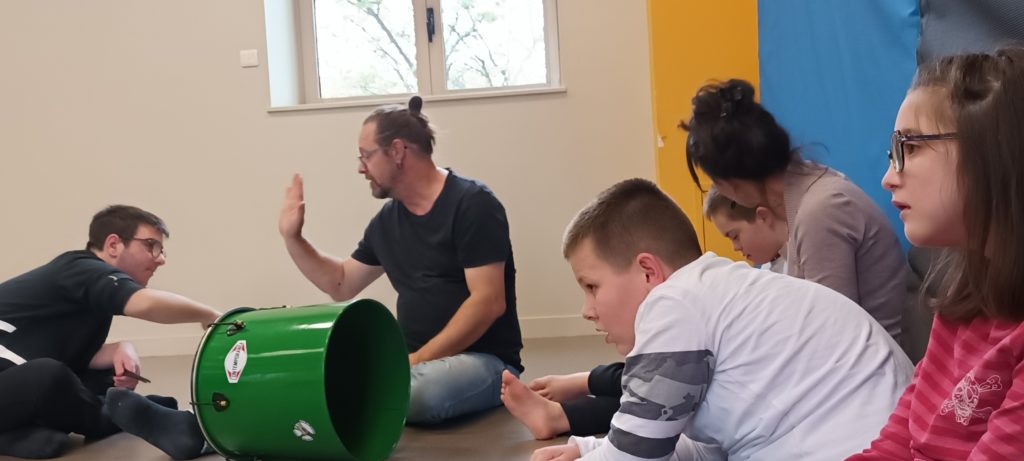 Culture et santé : À l’IME La Côtière, 3 jeunes préparent, avec deux animateurs des Zurbamateurs et de l’école de musique de Montluel, une batucada : ils sont assis dans une salle avec un tambour.