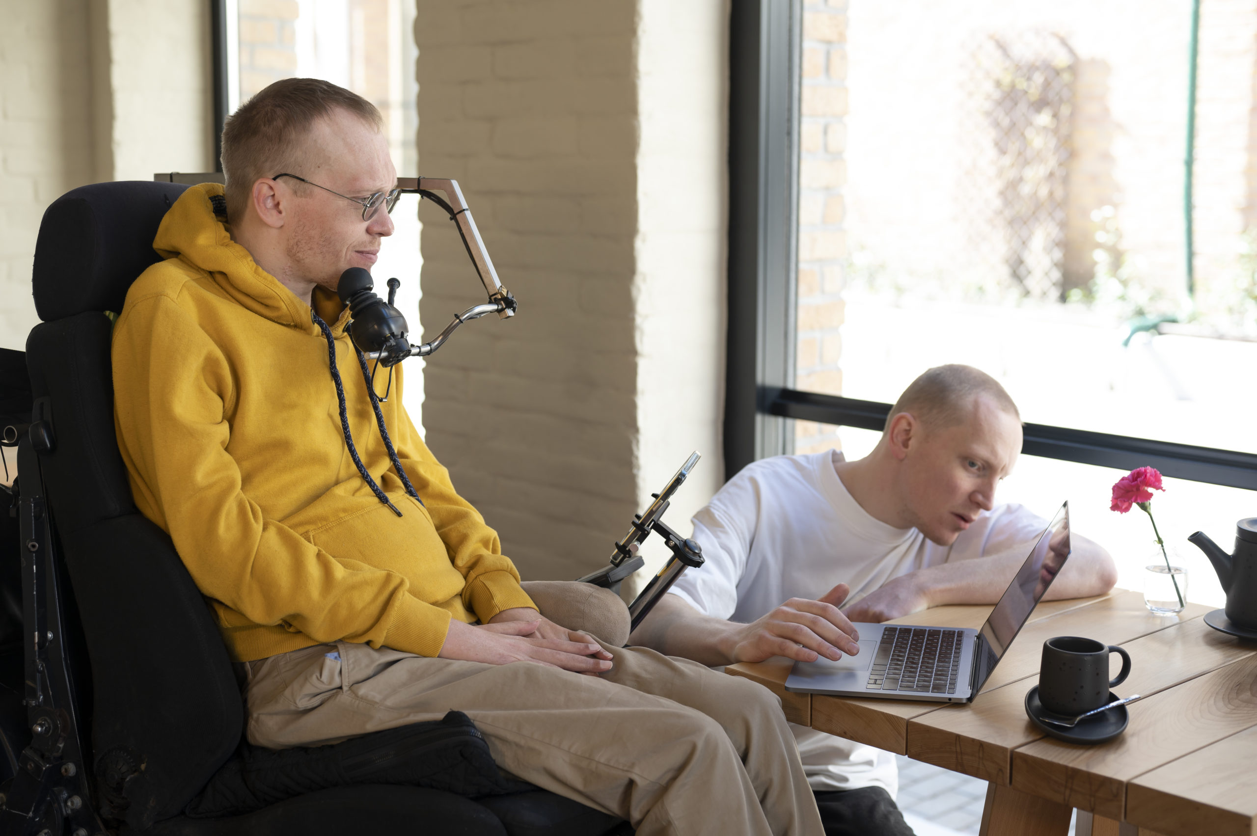 un homme en situation de handicap dans un fauteuil roulant est équipé d'un micro et d'un support d'écran. une autre personne l'aide à installer un ordinateur portable.